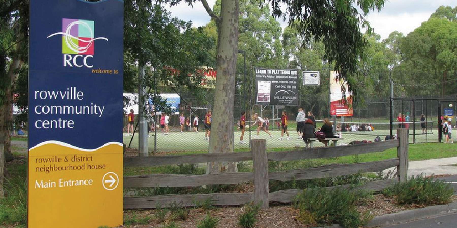 Rowville Community Centre sign in front of a lively recreation area with people engaging in outdoor sports and activities.