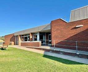 Image displays San Remo Recreation Centre building with brown bricks, concrete footpaths, a seat out the front on green grass