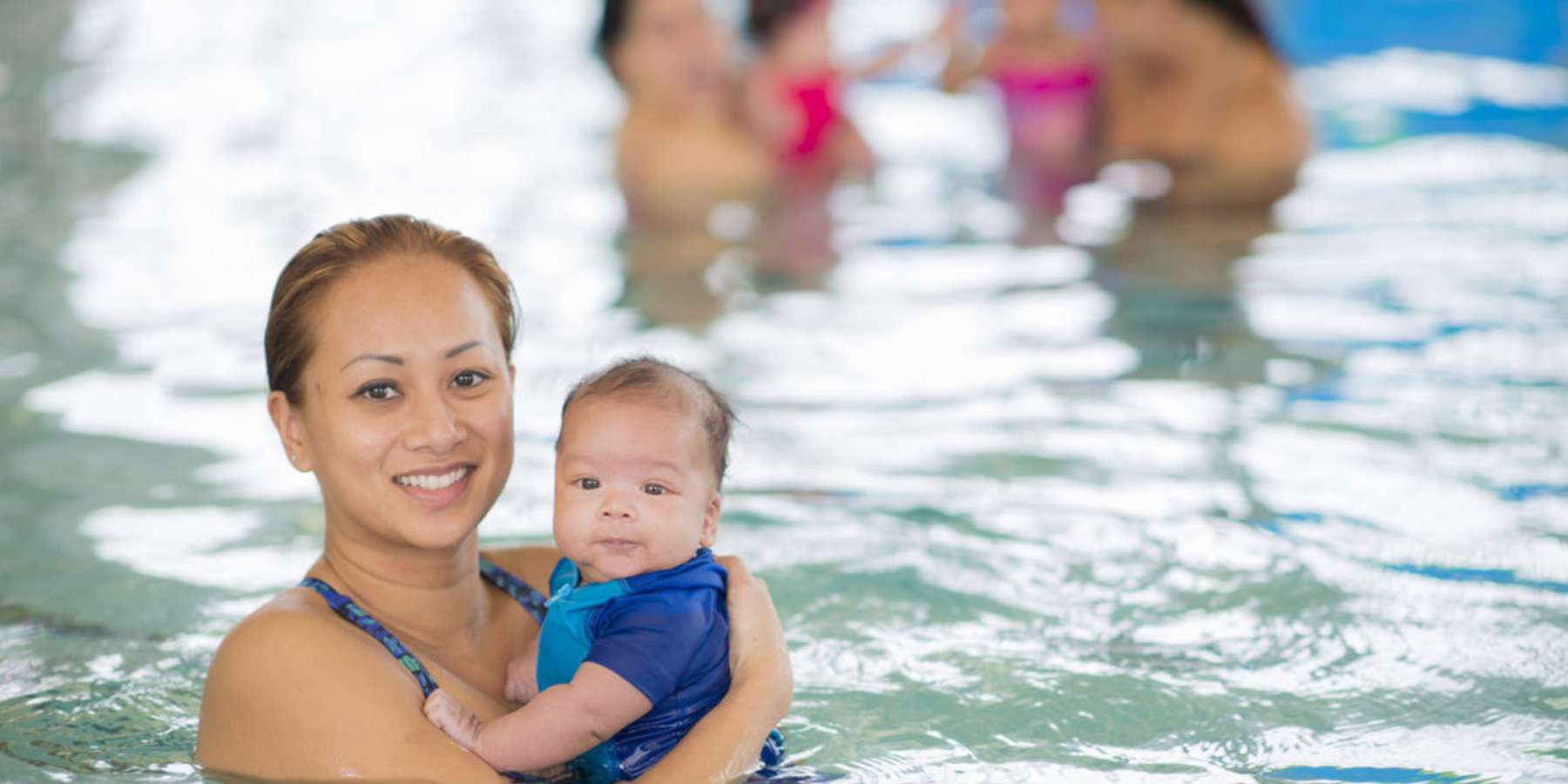 A caregiver holds a baby in a swimming pool, with other adults enjoying the water in the background. The scene is bright and cheerful.