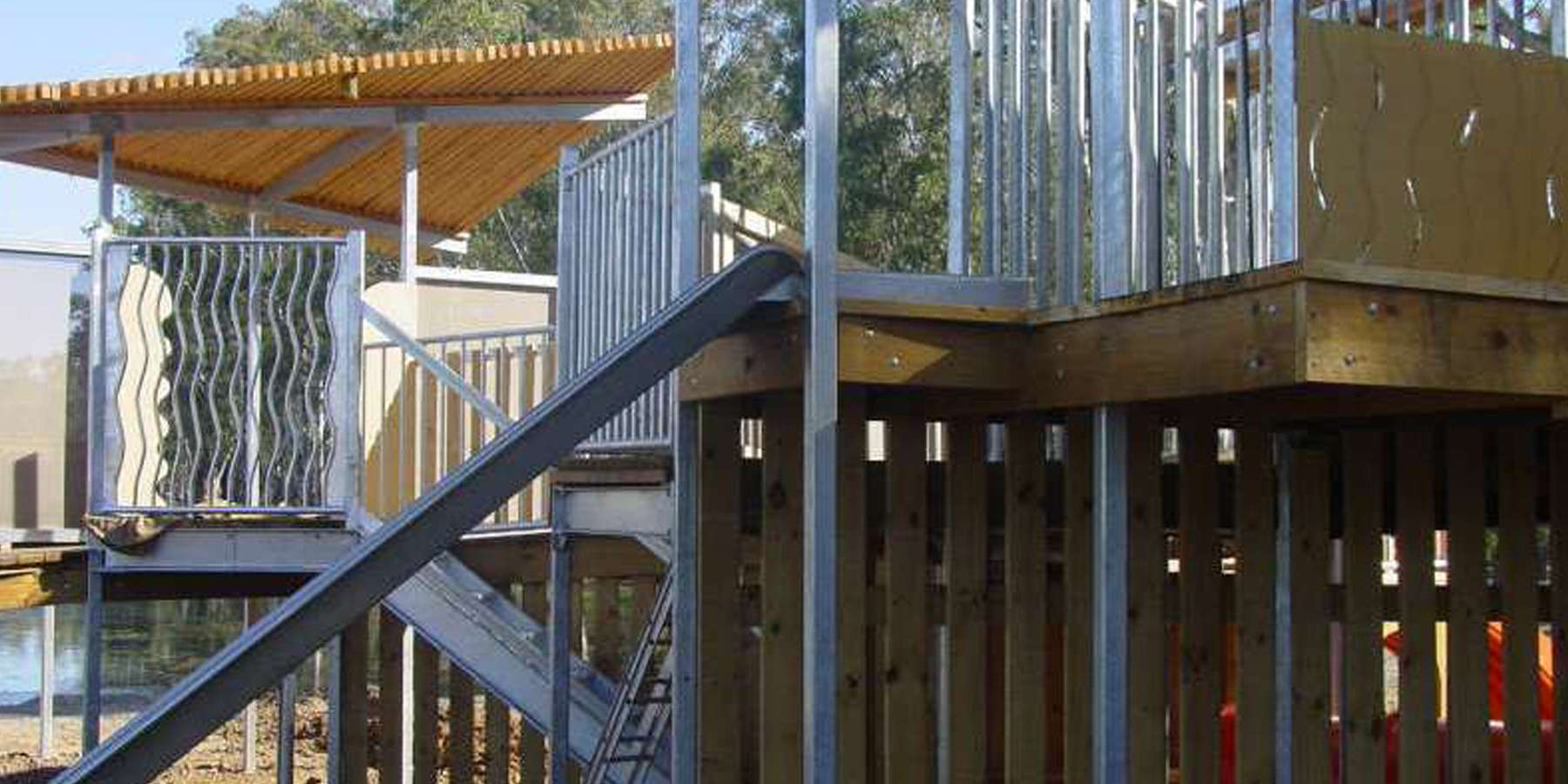 Close-up of a modern play structure featuring a staircase, wooden decks, and wavy metal barriers under a shaded canopy.