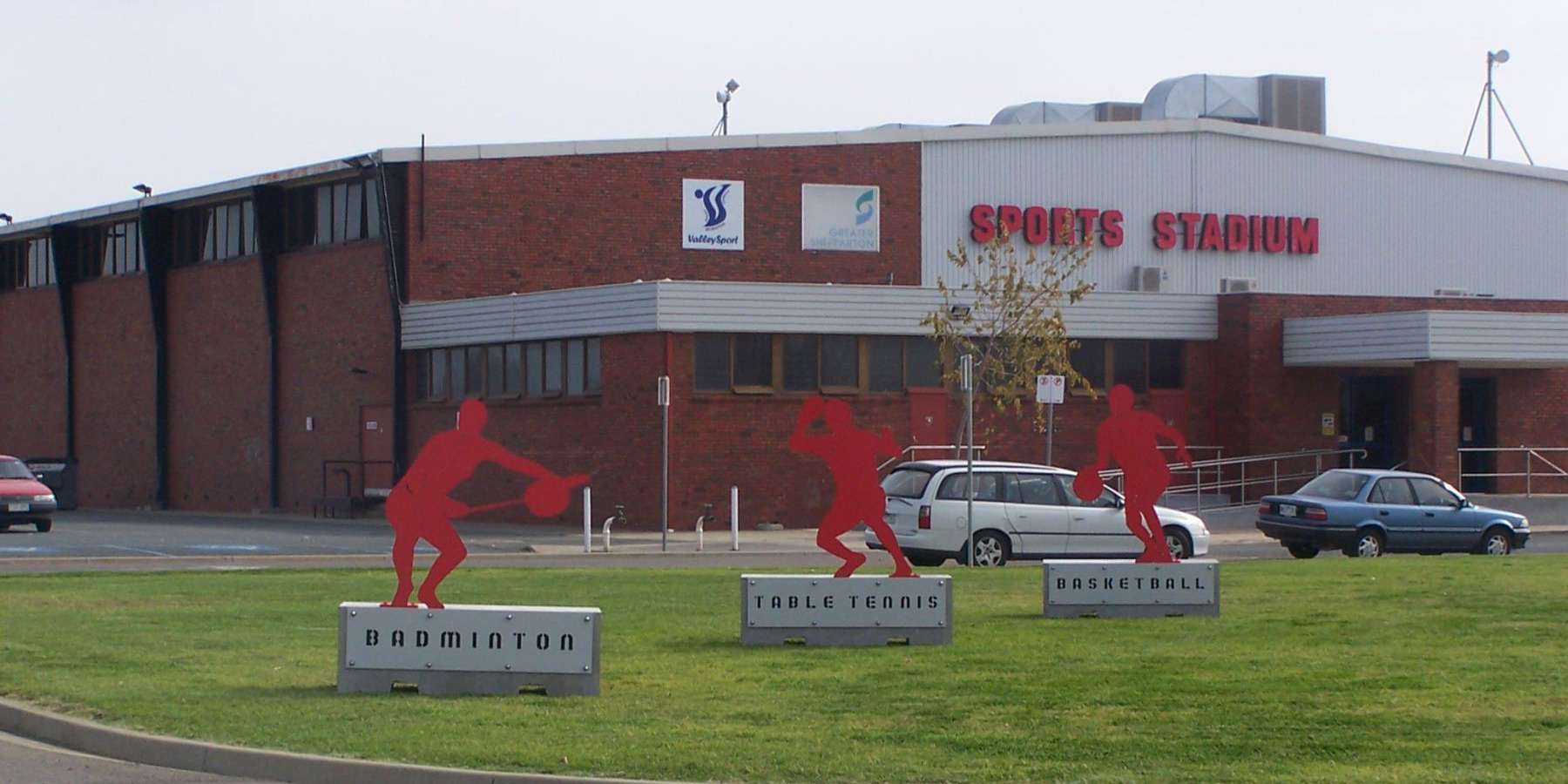 Exterior of a sports training facility featuring three red athlete sculptures on a grassy area with cars parked nearby.