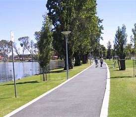 A scenic park pathway lined with trees beside a calm lake, where two people walk leisurely on a sunny day.