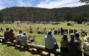 A crowd watches a horse riding event in a grassy park, surrounded by trees and parked vehicles under a clear blue sky.