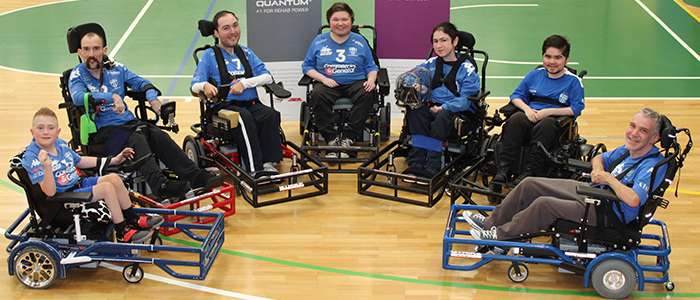 A group of six individuals in electric wheelchairs, smiling and dressed in matching blue shirts, gathered on a sports court.