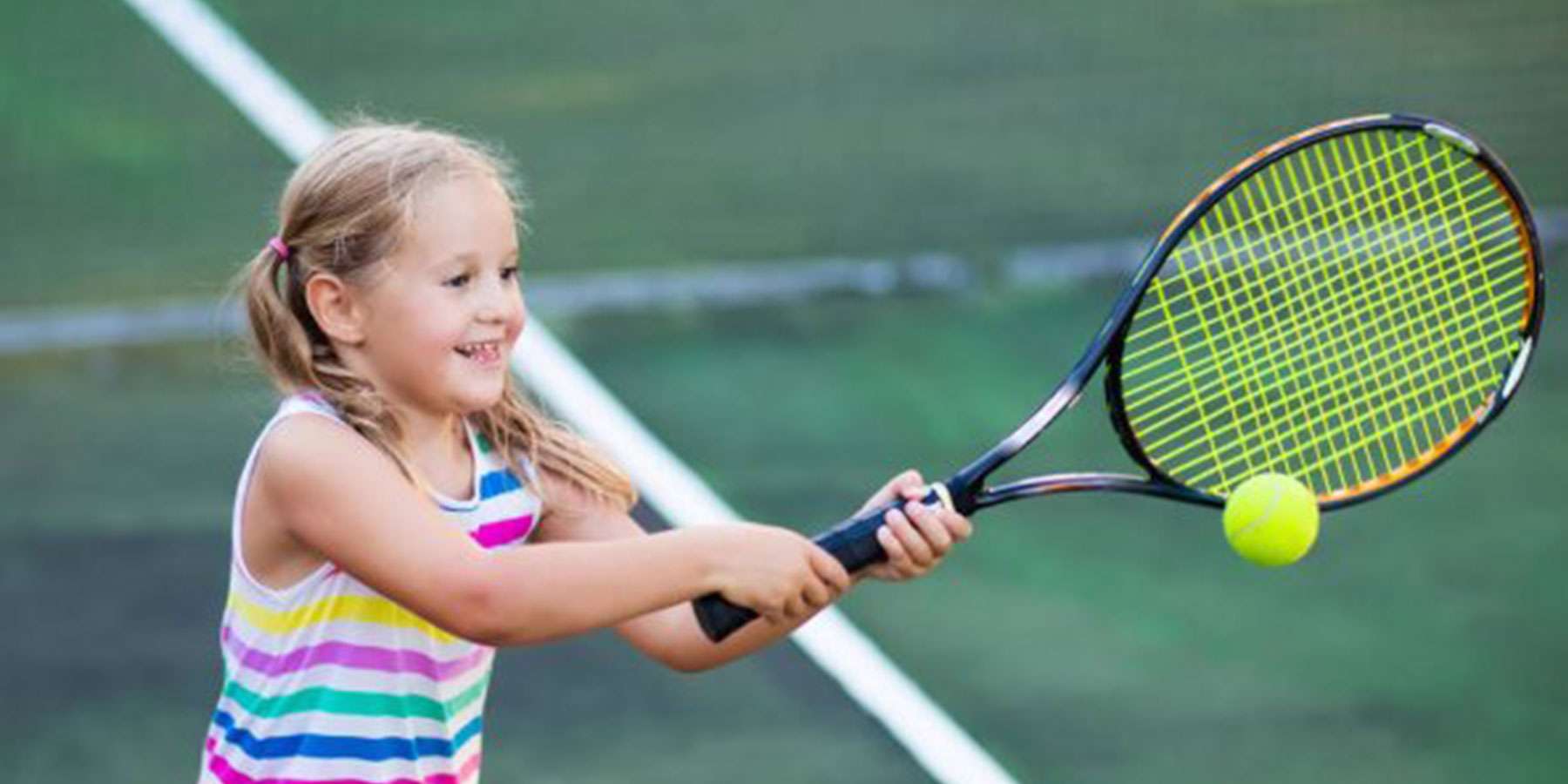 A child in a striped tank top swings a tennis racket at a bright yellow ball on a green court.