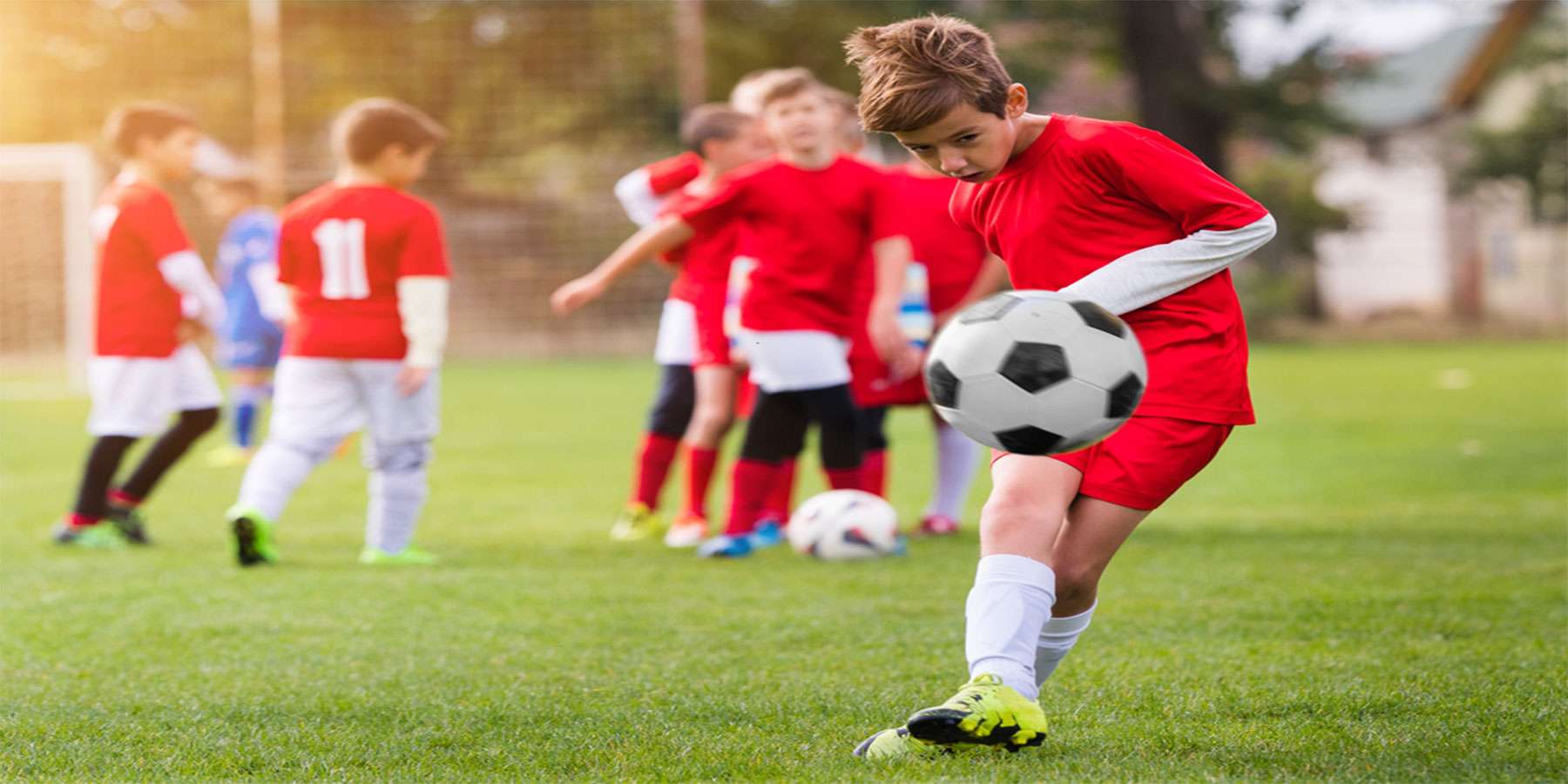 A young boy in a red jersey practices dribbling a soccer ball on a grassy field, with other players training in the background.