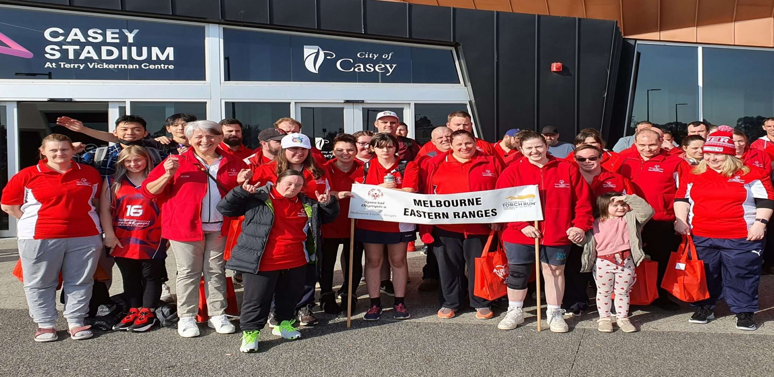 A group of people in red shirts stands outside Casey Stadium, holding a banner and smiling for the camera.
