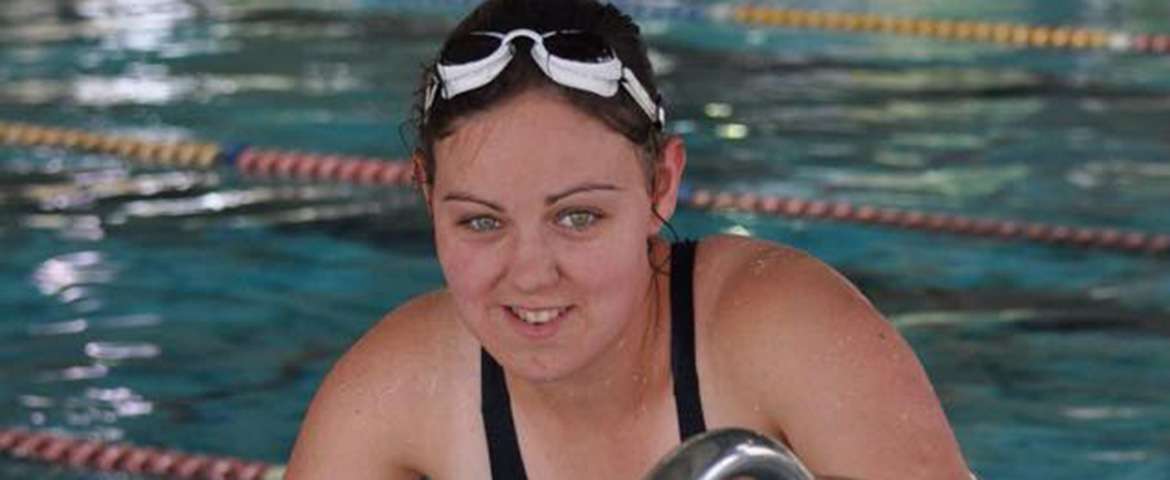 A swimmer wearing goggles and a swim cap poses at the edge of a pool, water visible in the background.