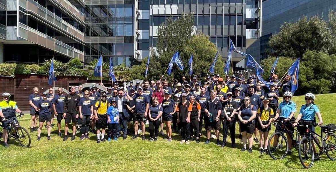 A large group of cyclists in matching shirts and helmets poses together on a sunny day outside modern buildings, holding blue flags.