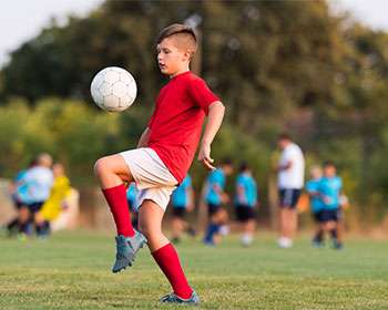 A young boy in a red jersey practices dribbling a soccer ball on a grassy field, with other players training in the background.