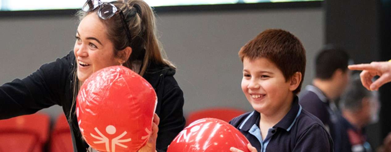A person assists a child with red sports equipment in a lively indoor setting, focusing on teamwork and activity.