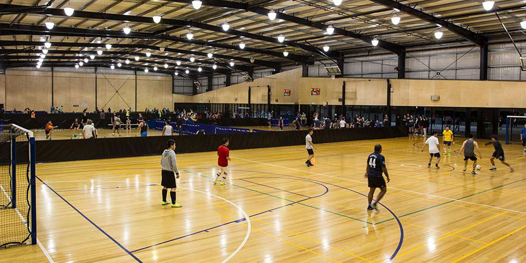 A spacious indoor sports facility with players engaged in a match on a wooden court, surrounded by spectators in the stands.