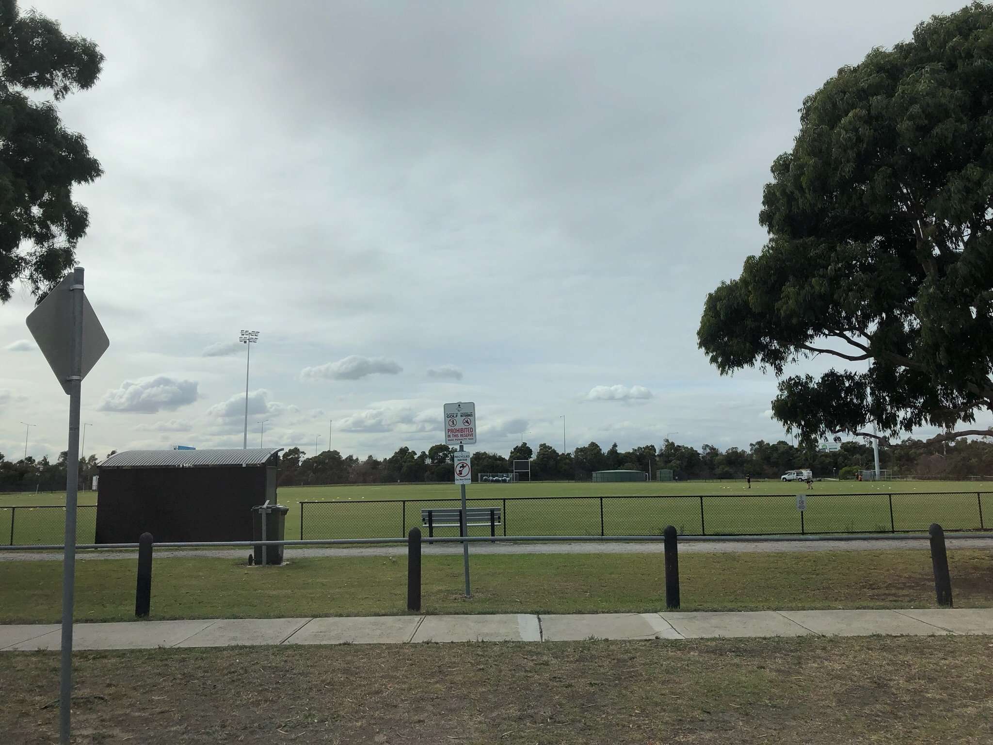 Image displays a green oval with a path, a fence and trees in the background. The sky is grey and overcast in this image