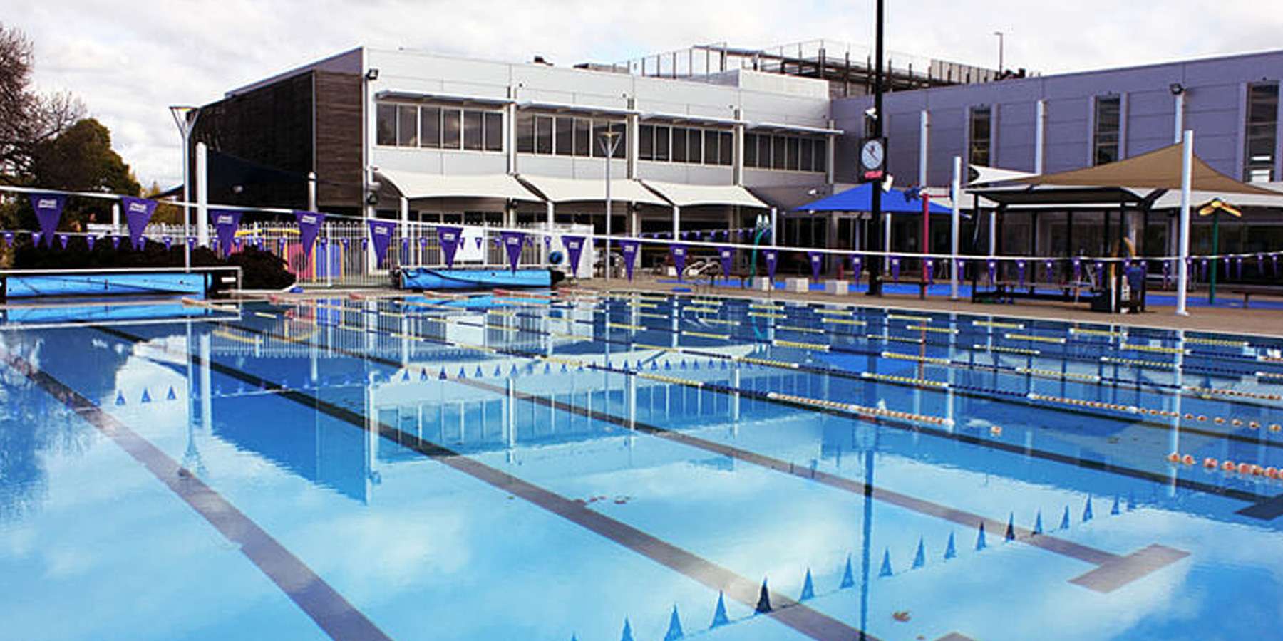 A sunny outdoor swimming pool with clear blue water, surrounded by lanes and a modern building in the background.