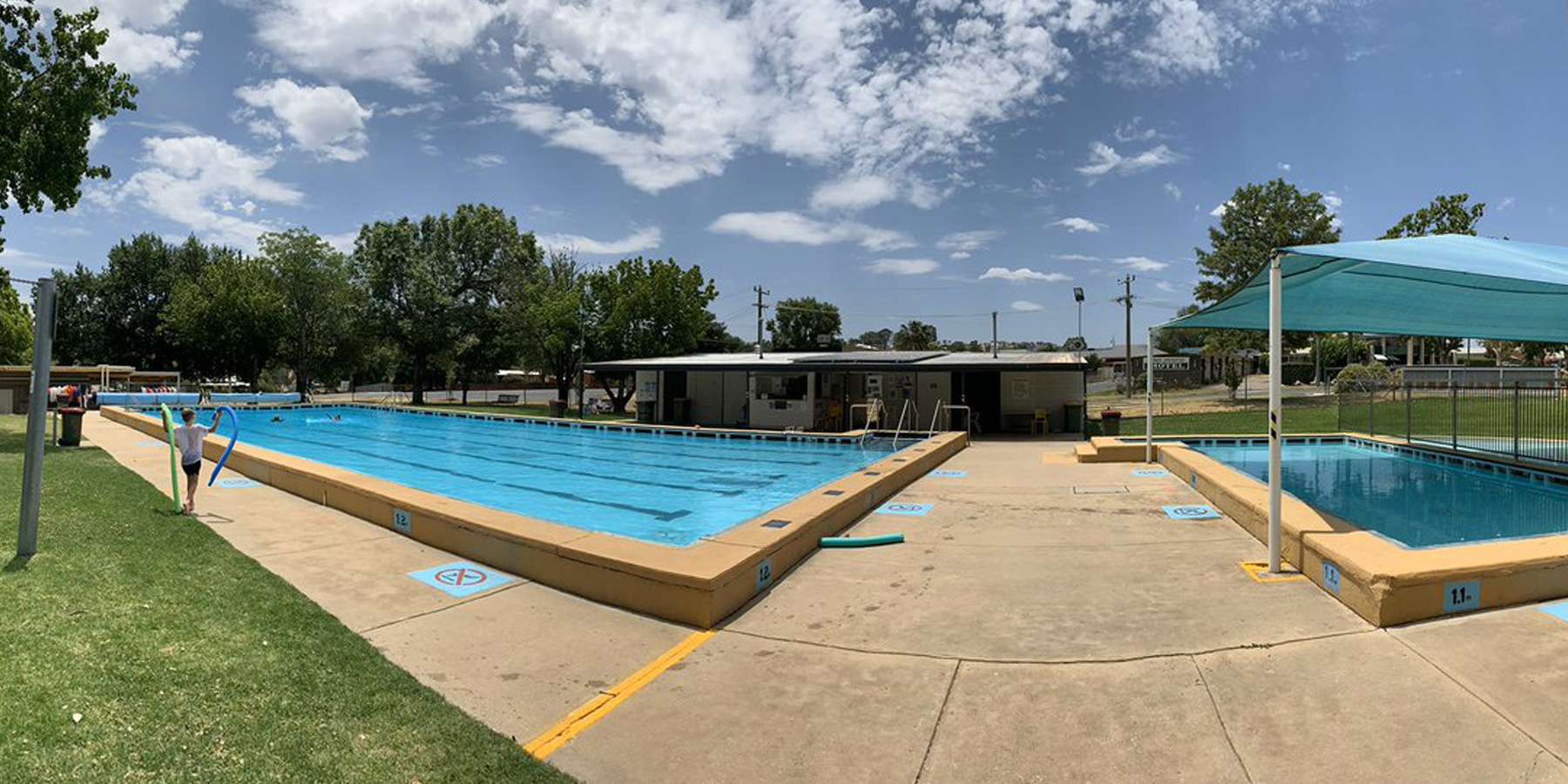 A sunny outdoor swimming pool area featuring two pools, surrounded by grassy lawns and trees under a partially cloudy sky.