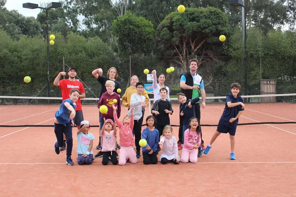 A group of children on a tennis court with red surface throwing tennis balls into the air.