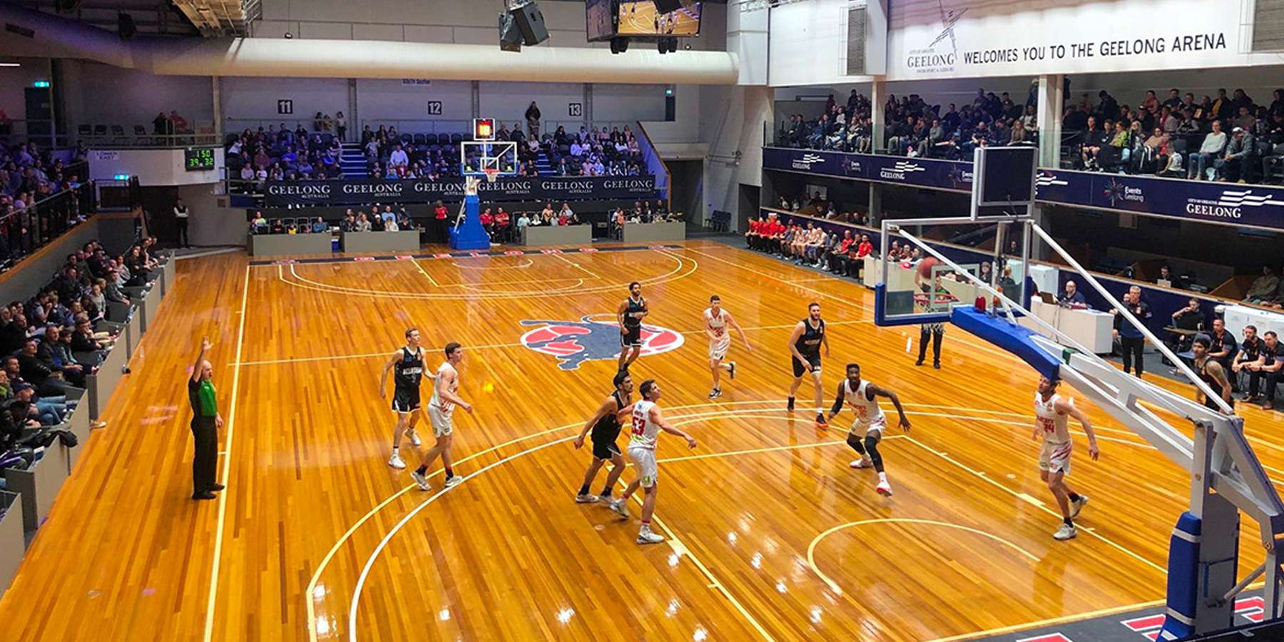 A basketball game in progress on a polished wooden court, with players in action and an audience in the stands.