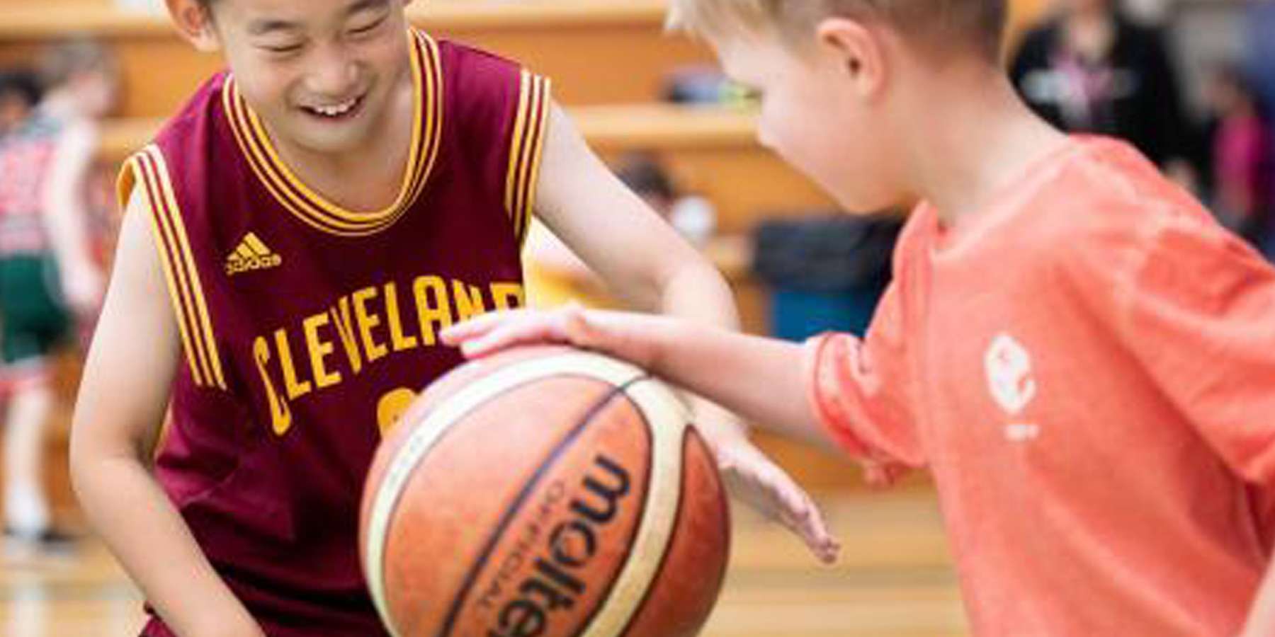 Two children play basketball, smiling and reaching for the ball.