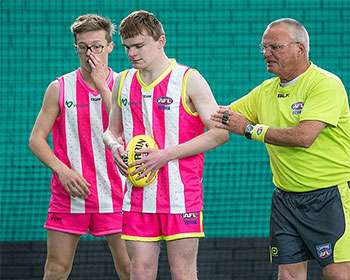 Two young athletes in pink and white jerseys stand with a referee, engaging in conversation on the field. A football is held in one player's hand.
