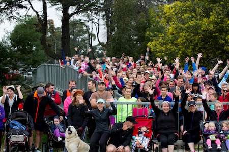 A large crowd of people cheer enthusiastically, raising their hands in the air, with strollers and a dog featured in the foreground.