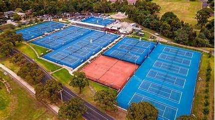 Image displays tennis multiple tennis courts with blue astro turf surfaces. There are trees way off in the distance.