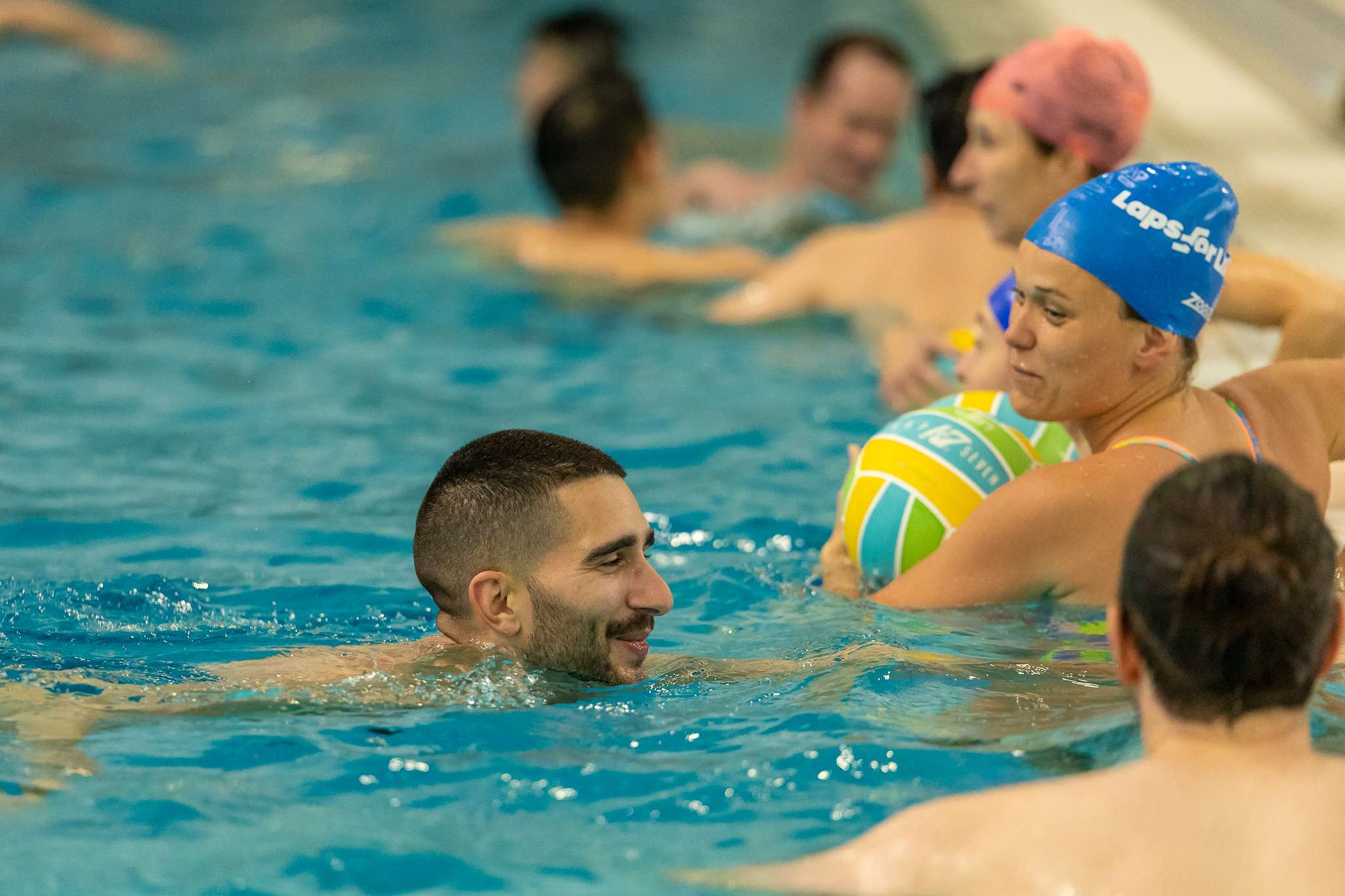 Photo of swimming pool with men and women in the water. One woman wearing a blue swimming cap is holding the ball.