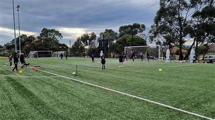 A soccer practice session on a lush green field with players in black jerseys, goalposts in the background, and a cloudy sky above.