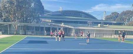 A sunny tennis court with players practicing, surrounded by trees and a modern sports facility in the background.