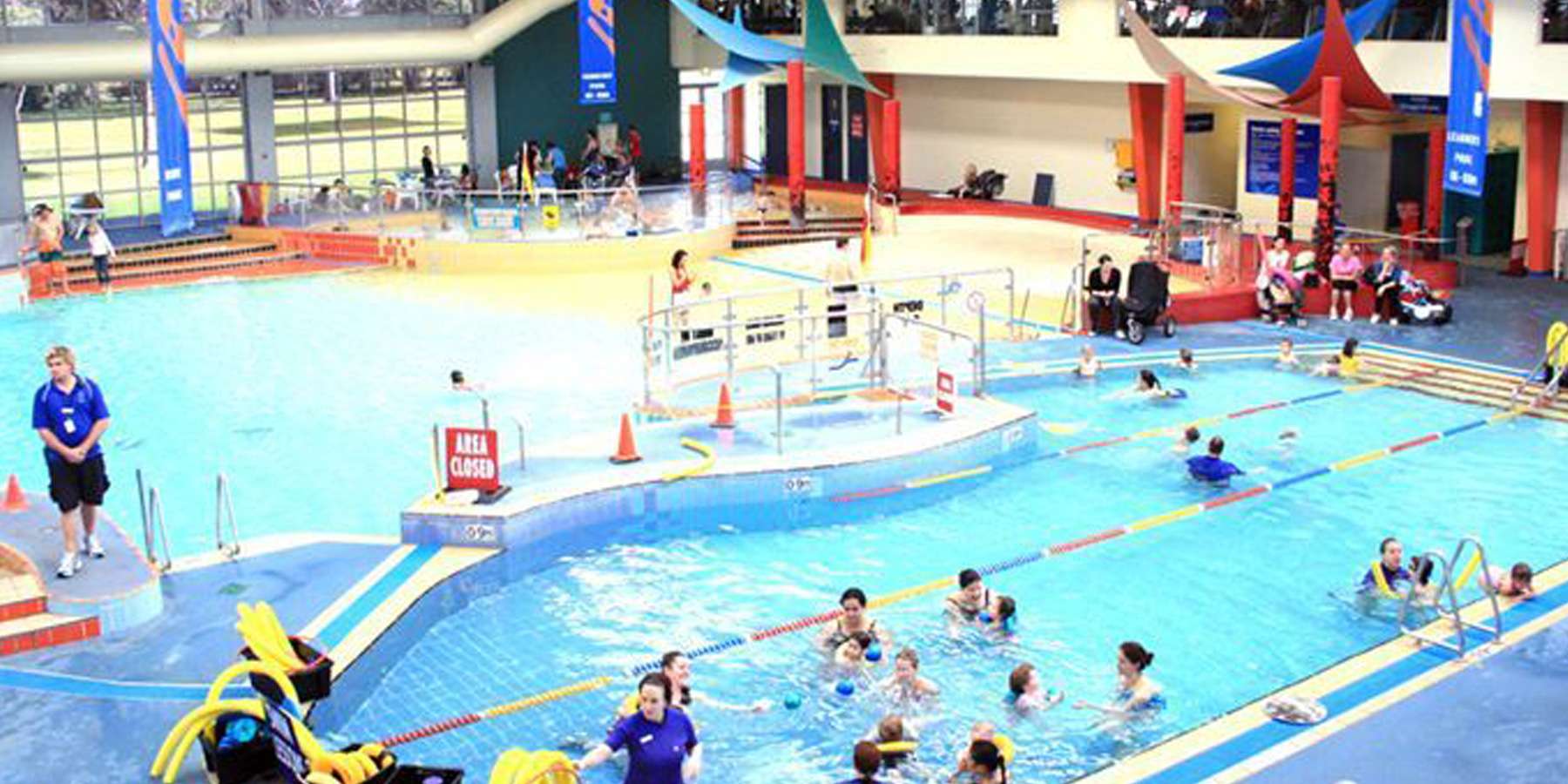 A vibrant indoor swimming pool filled with children playing, lifeguards on duty, and colorful decorations enhancing the lively atmosphere.