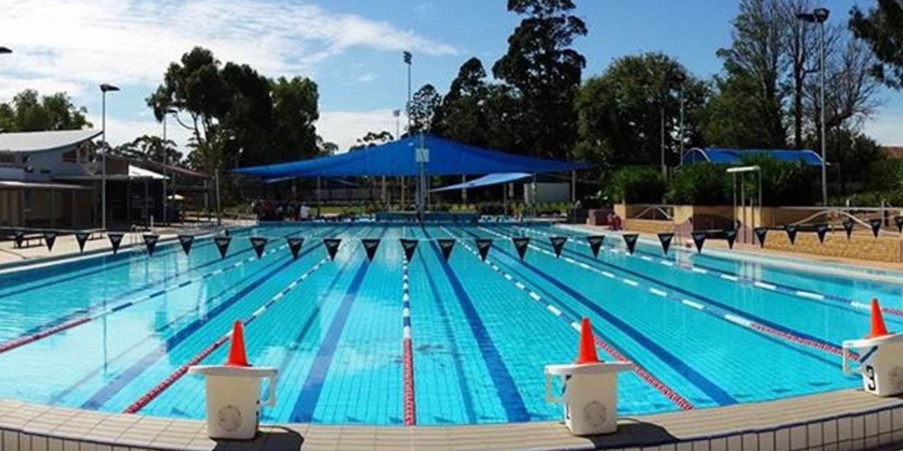 A clear, outdoor swimming pool with lane markers and red cones, shaded by blue canopies, surrounded by green trees under a bright sky.