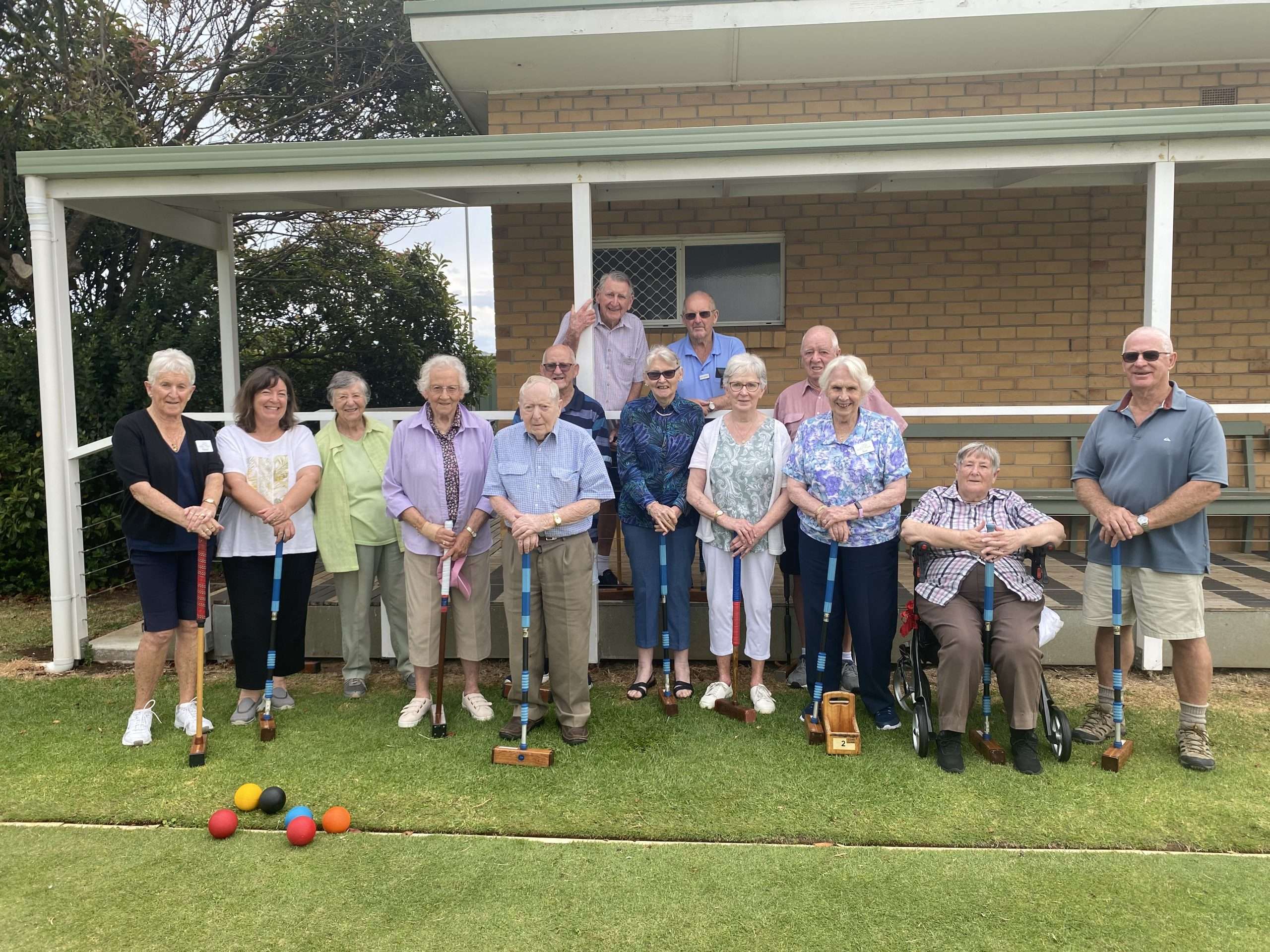A group of seniors standing on a grassy lawn, holding croquet mallets, with colorful balls placed in front of them.