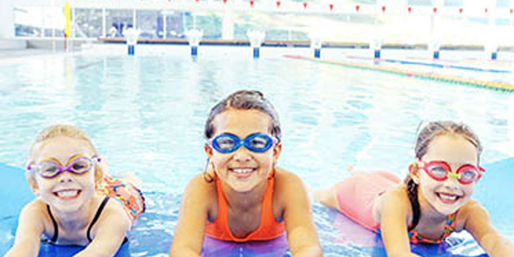 Three children in colorful swimsuits lie on floating mats in a bright, indoor swimming pool, enjoying a fun day in the water.