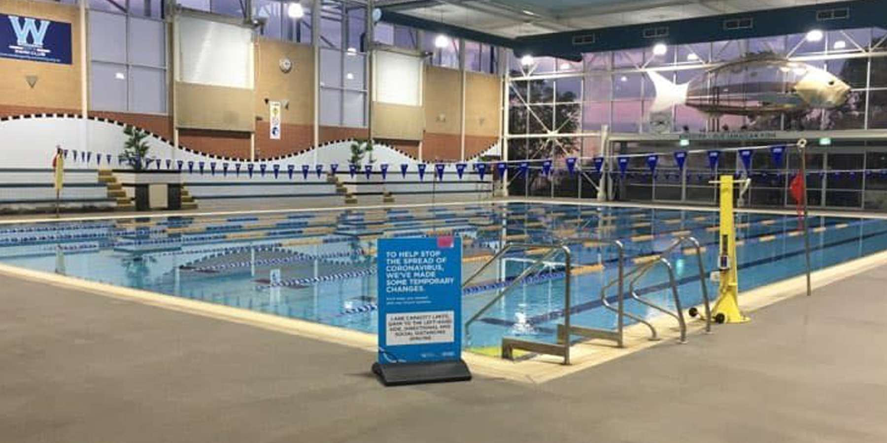 Indoor swimming pool with lane markers, a safety sign, and large windows allowing natural light in.