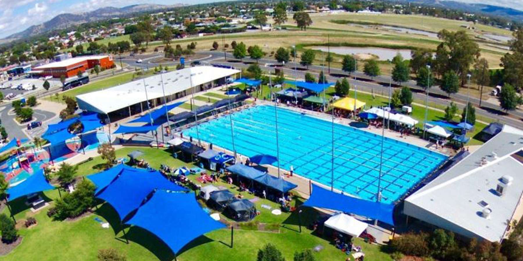 Aerial view of a large swimming pool complex surrounded by blue tents, lush grass, and distant hills, bustling with activity.