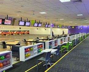 Image displays a brightly lit bowling alley, with many lanes. There are shelves with all different colour bowling balls, and televisions above each lane. The carpet is grey and there are small chairs and tables near each lane.