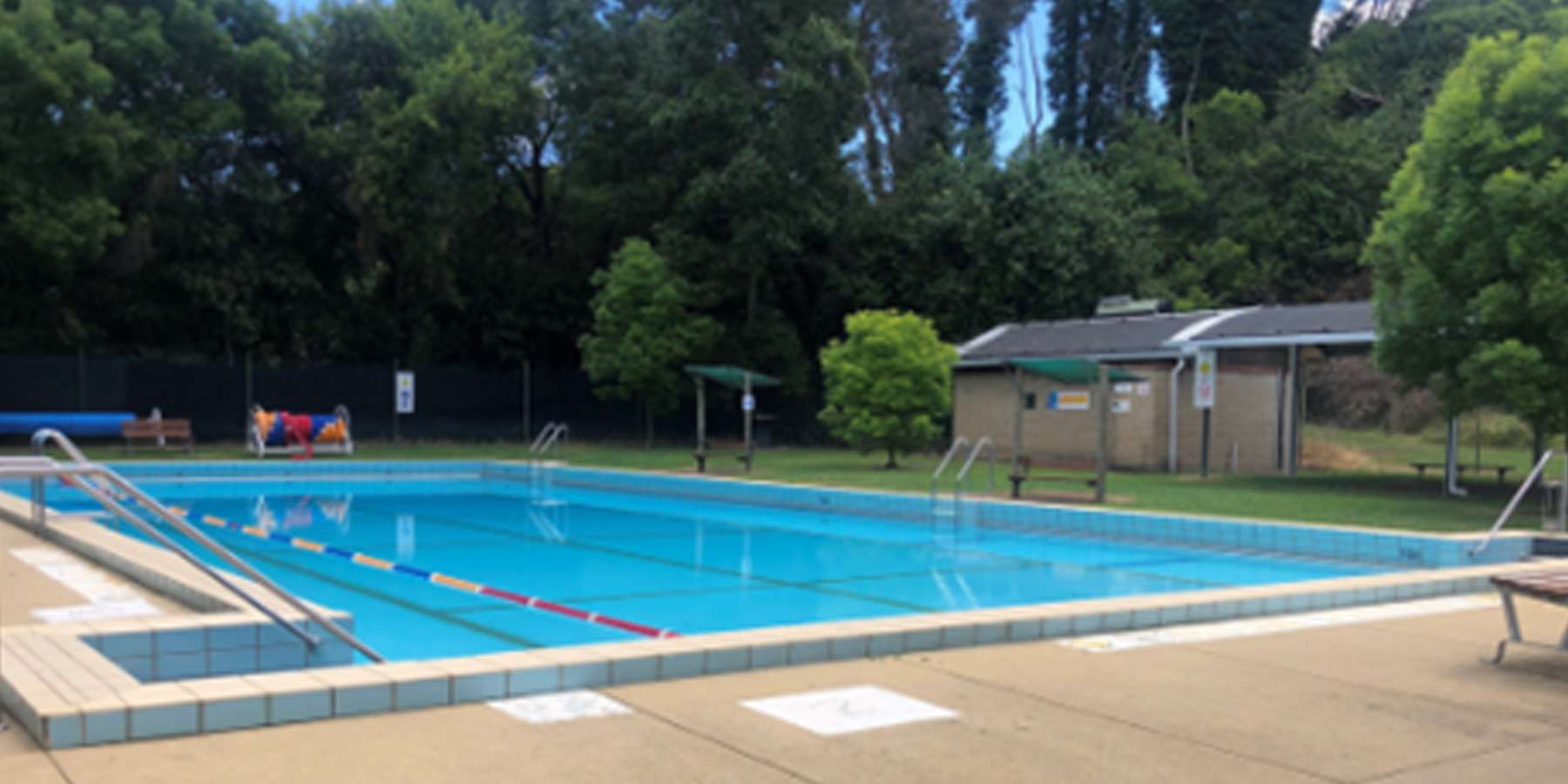A clear blue swimming pool surrounded by a concrete deck, with green trees and a building in the background under bright sunlight.