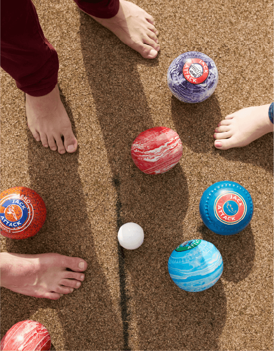Feet stand on sandy ground surrounded by colorful bowls balls, with a smaller white ball at the center of the scene.