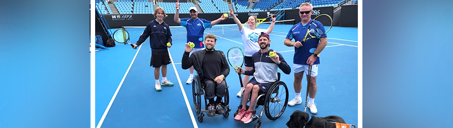 A group of six people, including two in wheelchairs, pose joyfully with tennis rackets on a blue court, accompanied by a black dog.