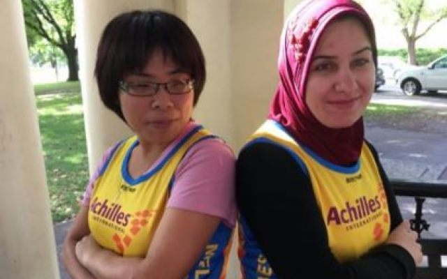 Two women standing back-to-back outside, wearing colorful athletic jerseys. The setting features green grass and parked cars.