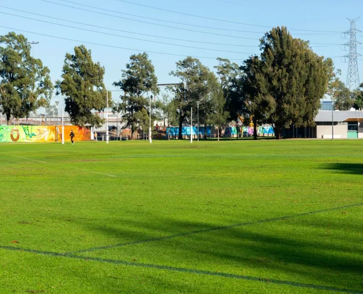 Vibrant green sports field with trees and colorful murals in the background under a clear blue sky, with one person jogging.