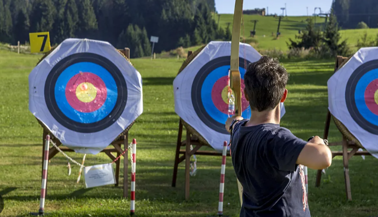 A young archer stands in a field, aiming at colorful target boards, with green hills in the background and blue skies above.