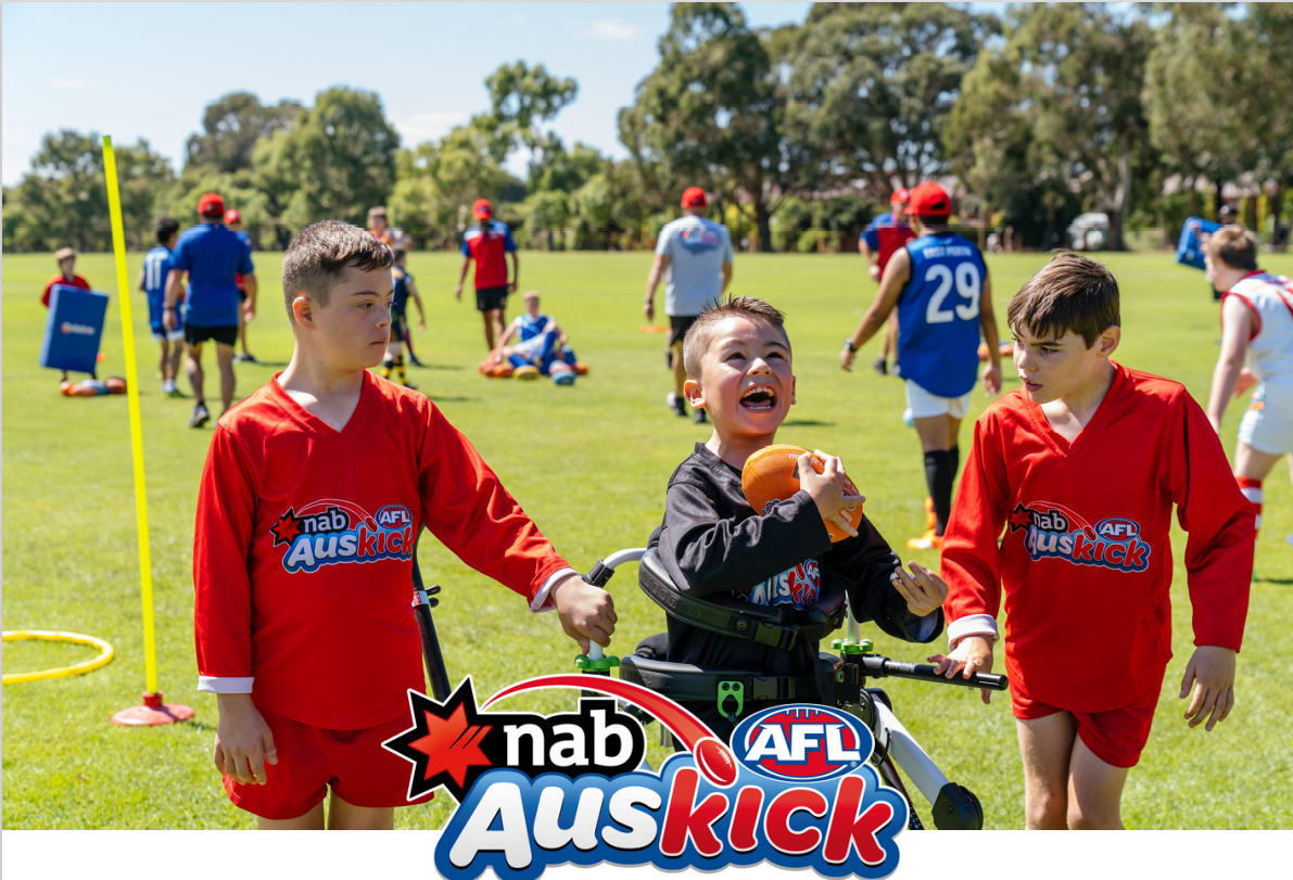 Two children in red jerseys interact with a boy in a wheelchair, while other kids engage in AFL activities on a sunny outdoor field.