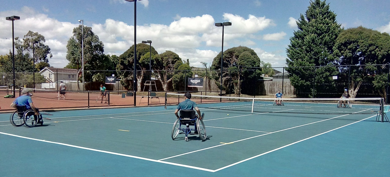 Two wheelchair tennis players on a blue court, with trees and cloudy skies in the background. Sports equipment is visible nearby.
