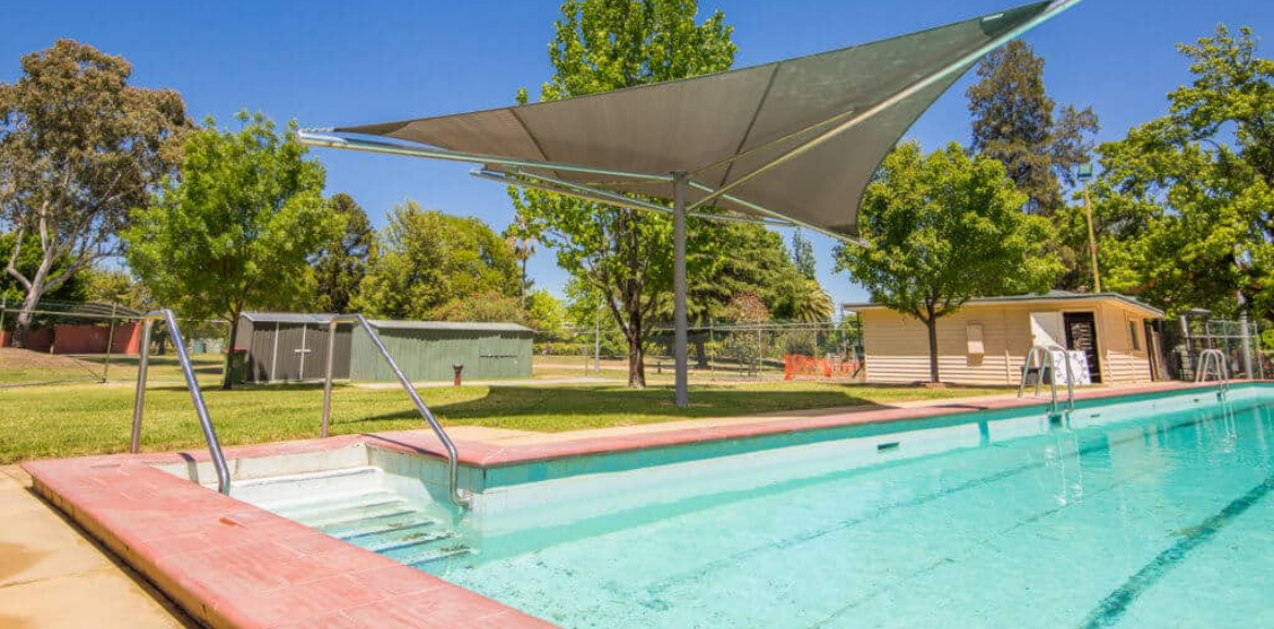 A large inground ppol surrounded by green trees and grass and a cream brick changeroom in the background.