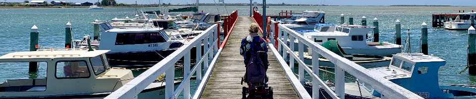A person on a mobility scooter ona pier with white railing and fishing boats on either side on blue water