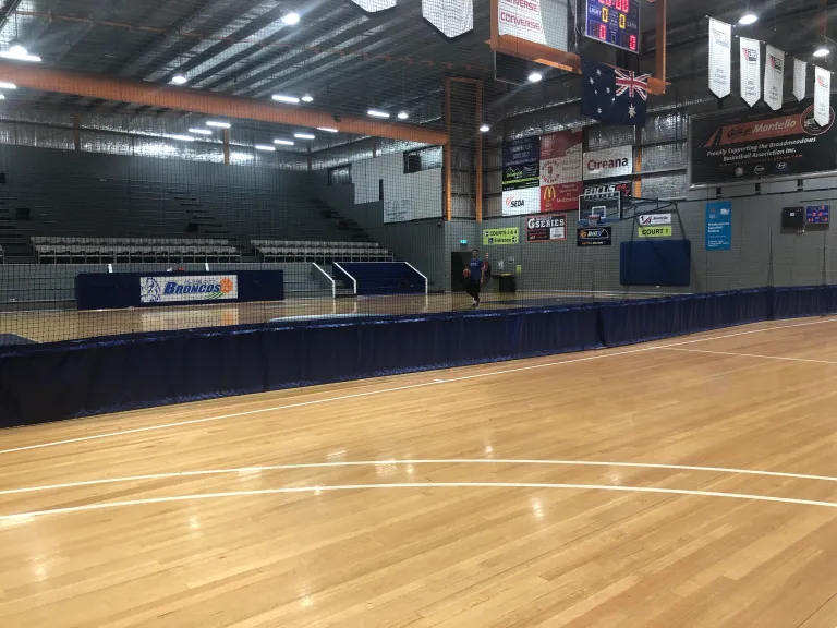 A spacious indoor basketball court with polished wooden flooring, empty bleachers, and banners hanging from the ceiling.