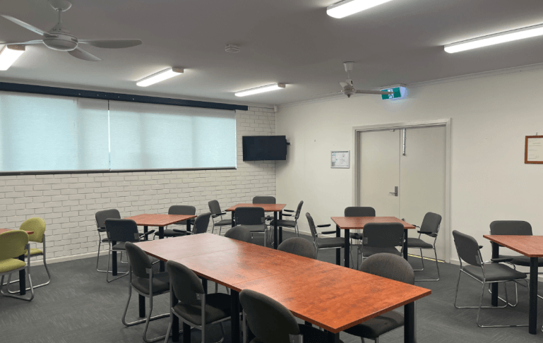 A bright meeting room with wooden tables, gray chairs, a wall-mounted TV, and large windows covered by light shades.