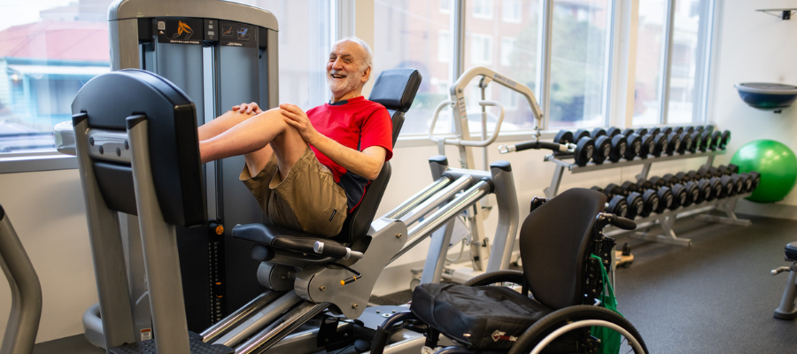 A man using gym equipment and smiling, with a wheelchair next to him