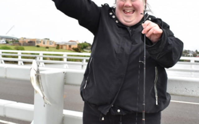 A smiling woman in a black jacket holds up a fish she caught while standing on a bridge, showcasing her excitement.