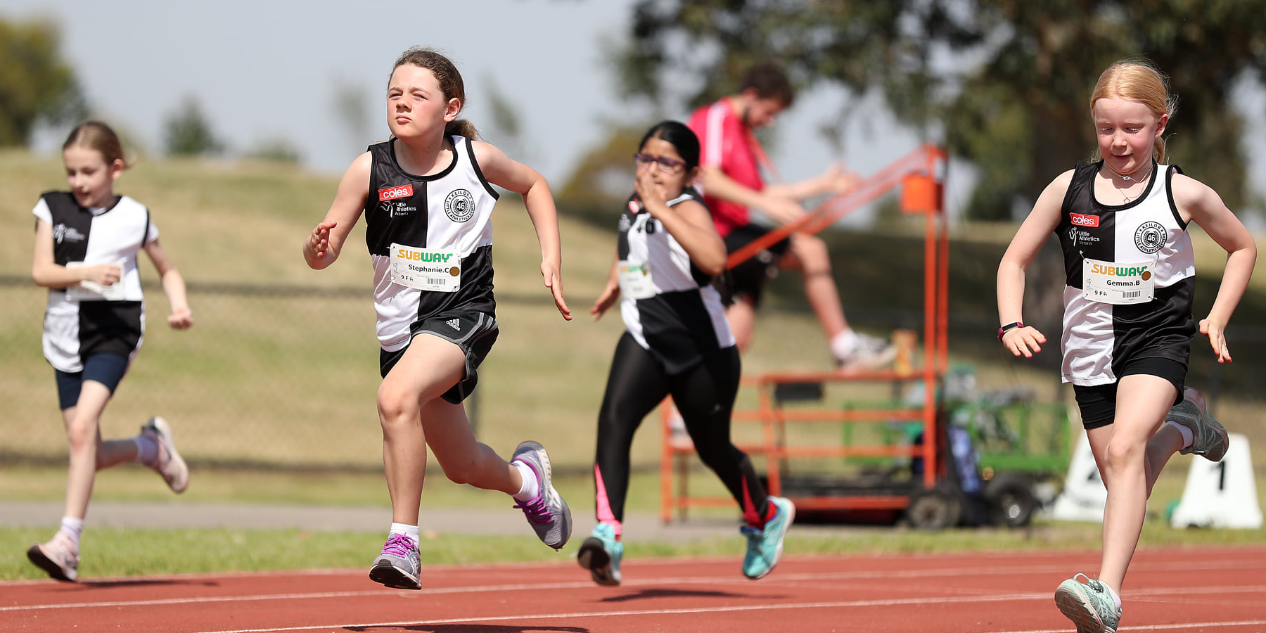 Four young girls in athletic gear sprint down a track, competing in a race, while a coach observes in the background.